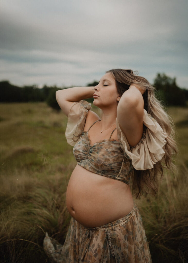 Pregnant woman standing in a field with her hands in her hair, wearing a soft floral gown during an outdoor maternity session with Raleigh maternity photographer.