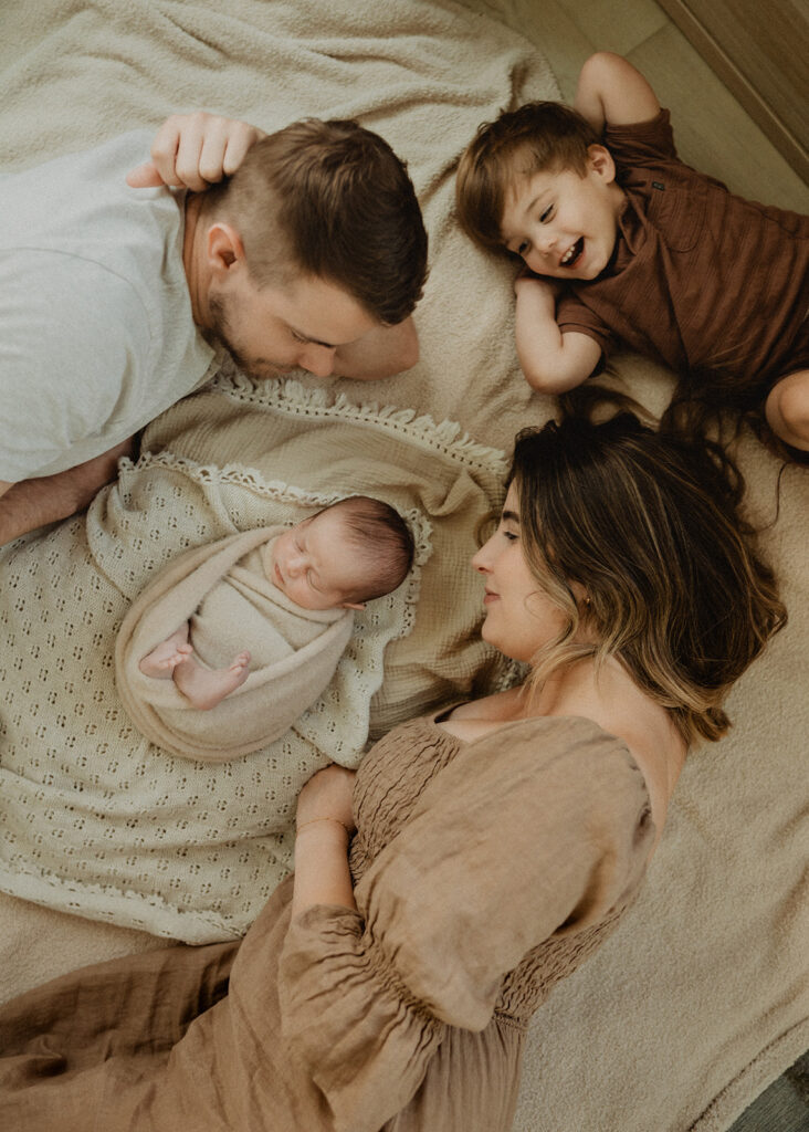 Overhead view of a family lying together on a soft blanket with their newborn swaddled in neutral tones, creating a peaceful lifestyle scene that represents family connection and preparation for infant swim lessons Durham NC.