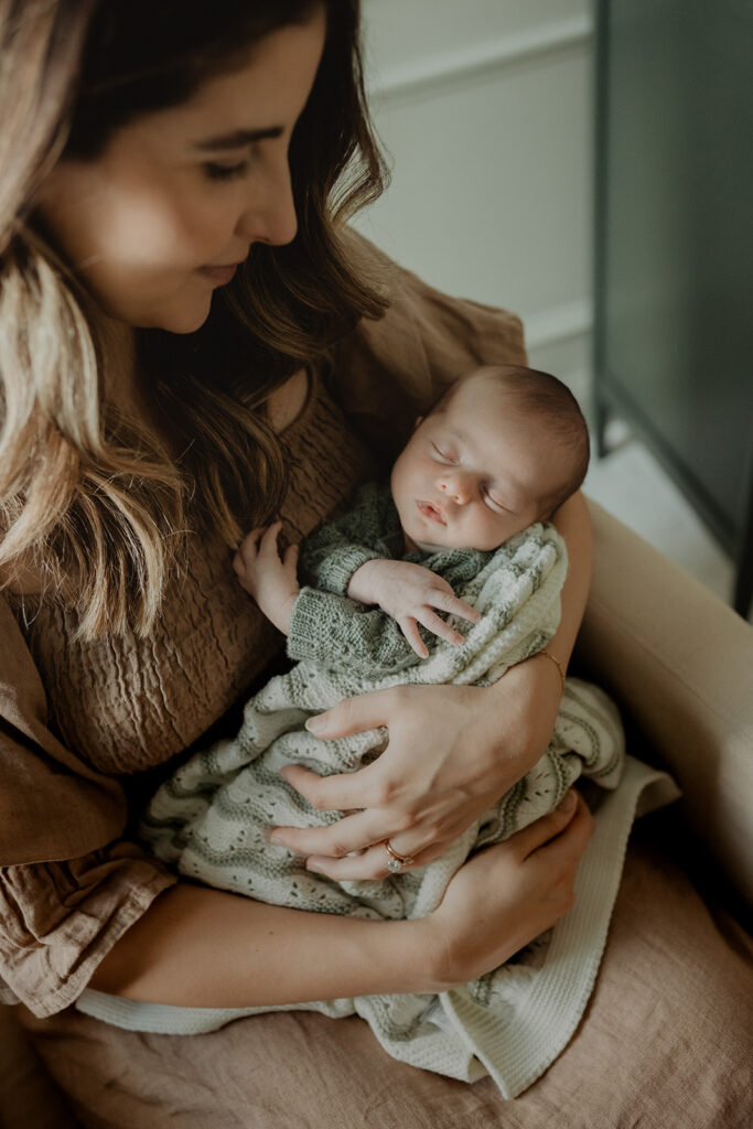 Close-up of a mother holding her sleeping newborn son near a window with soft natural light, highlighting calm early days of parenthood.