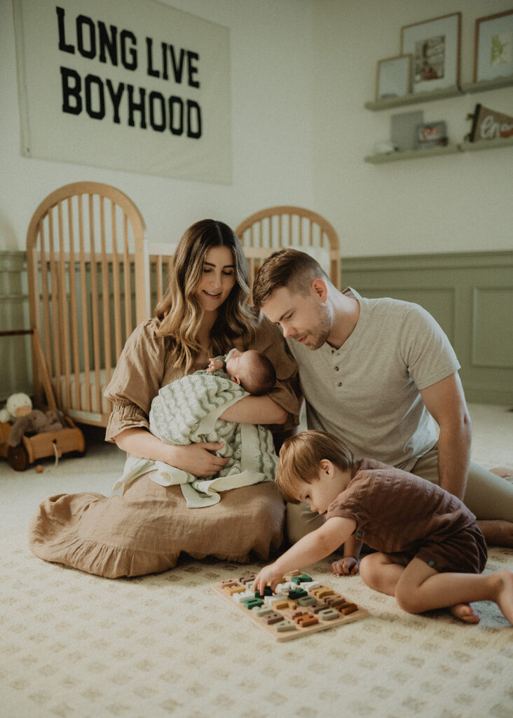 Family gathered in a nursery with a newborn and toddler playing on the floor, a cozy in-home lifestyle portrait showing daily family life before enrolling in infant swim lessons Durham NC.
