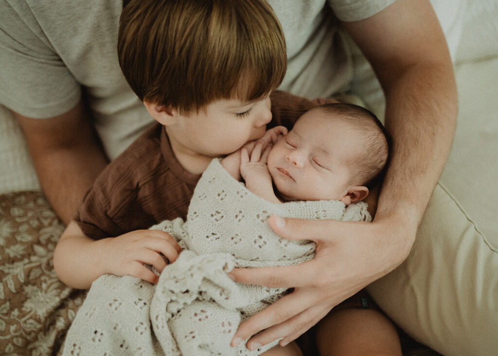 Toddler gently kissing his newborn baby brother while sitting in a parent’s arms, capturing sibling bonding and family milestones during the infant stage in Durham NC.