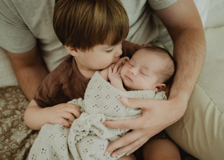 Toddler gently kissing his newborn baby brother while sitting in a parent’s arms, capturing sibling bonding and family milestones during the infant stage in Durham NC.
