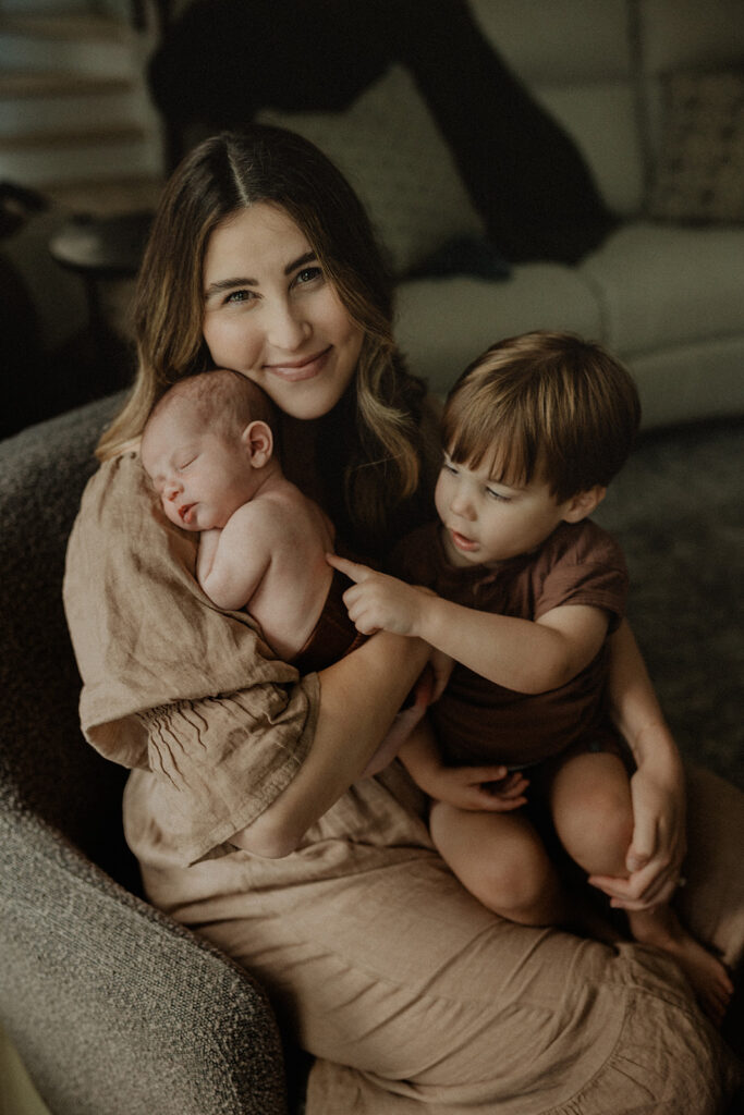 Mother sitting on a sofa holding her sleeping newborn while her toddler gently points at the baby, captured during an in-home family moment that reflects early bonding before starting infant swim lessons Durham NC.