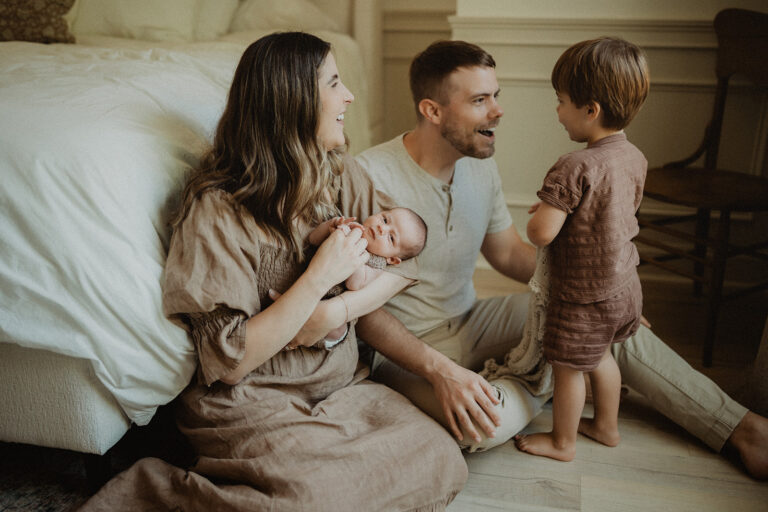 Family sitting on the floor beside the bed as parents interact with their toddler while holding their newborn. In-home photo session in Durham, NC.