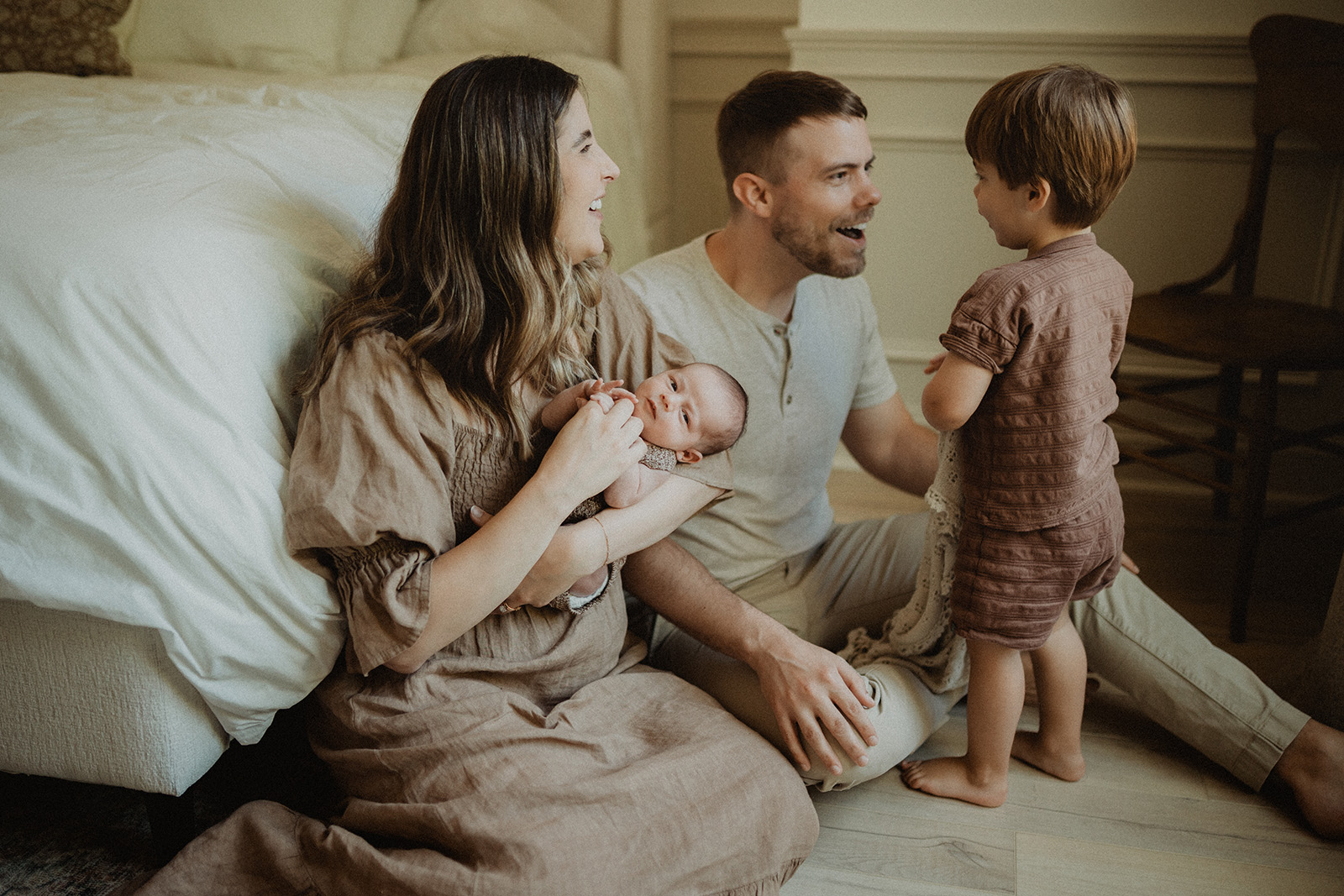 Family sitting on the floor beside the bed as parents interact with their toddler while holding their newborn. In-home photo session in Durham, NC.