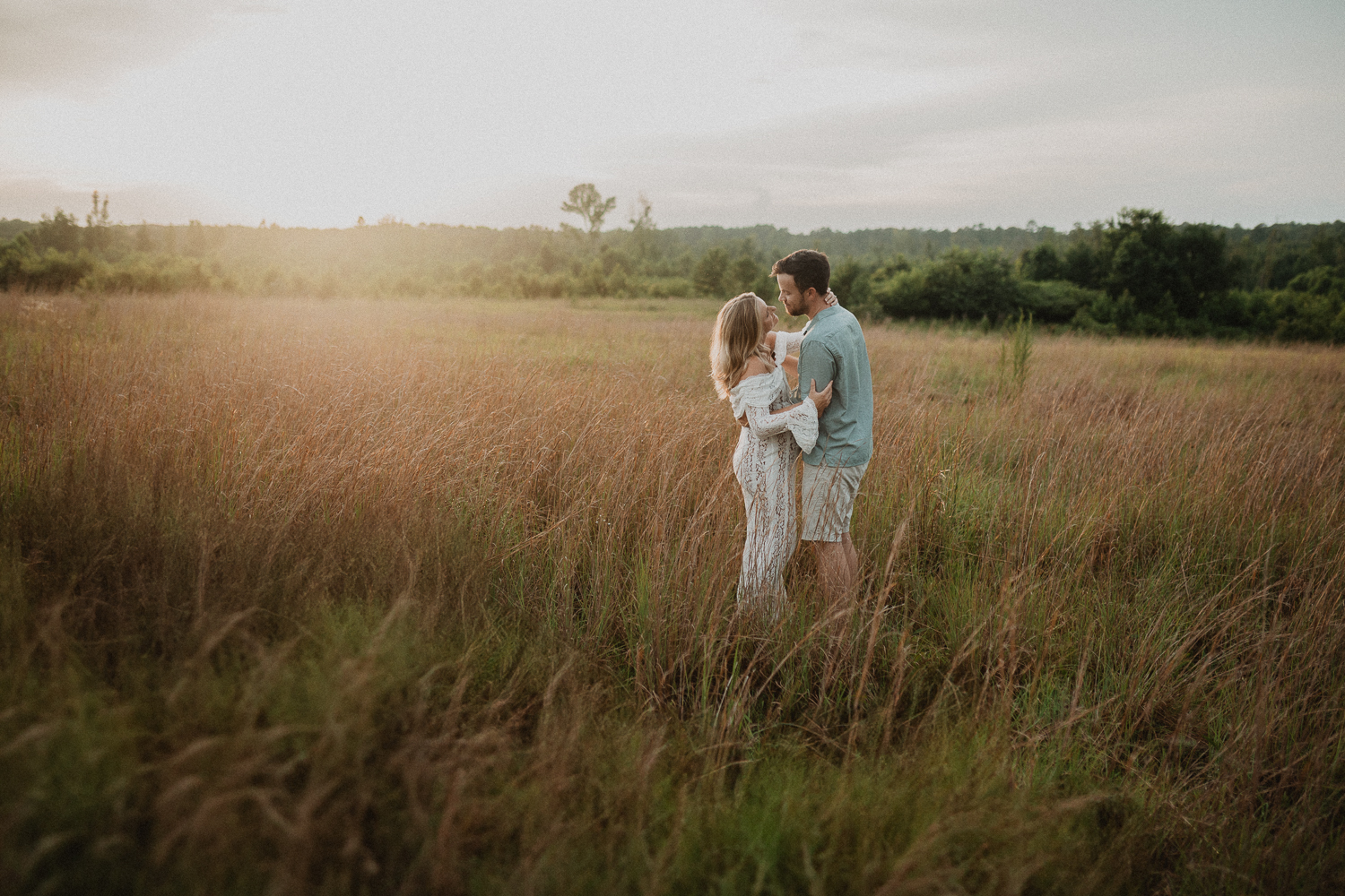 Scenic golden-hour maternity portrait showing an expecting couple standing embraced in the middle of a glowing field, surrounded by warm sunlight and tall grass. The image was taken near Lucky Elephant Yoga and Wellness by Victoria Vasilyeva Photography.