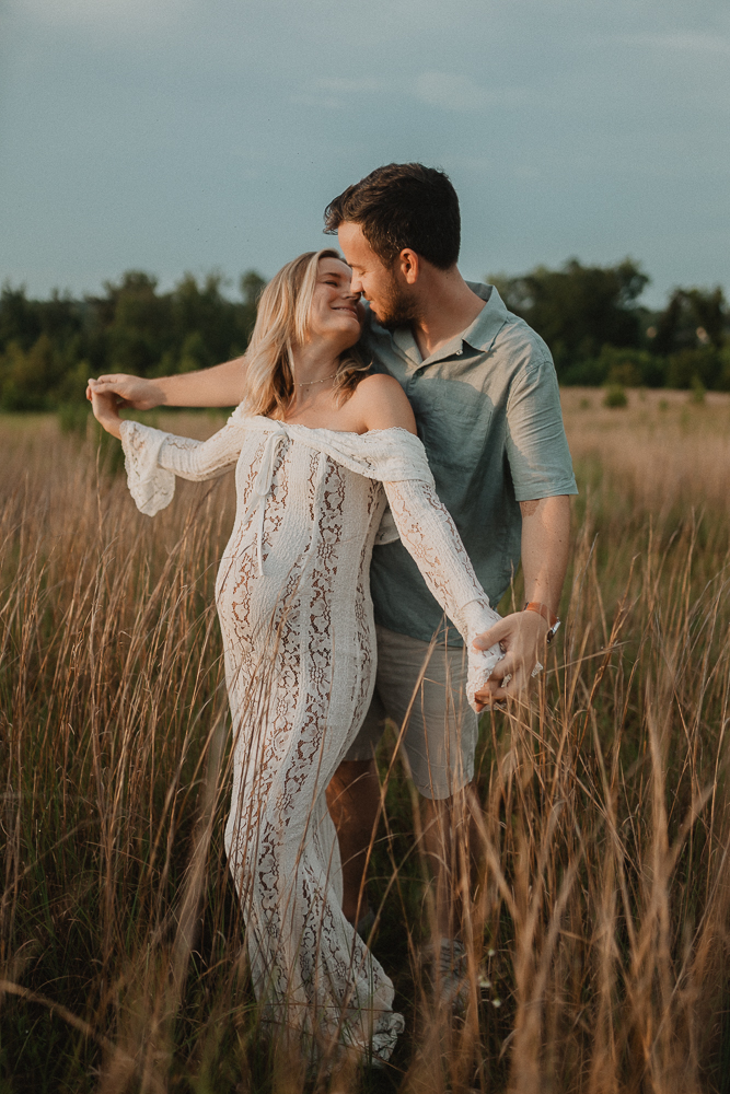 Playful maternity photo of a couple embracing in a tall grassy field at sunset near Lucky Elephant Yoga and Wellness. The expecting mother leaning back into her partner while smiling. Photo by maternity photographer - Victoria Vasilyeva Photography.