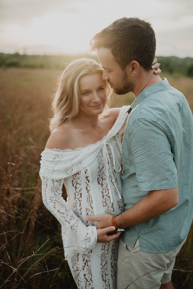 Close-up maternity portrait of a couple holding each other in a golden field, the expecting mother in a white lace dress and her partner in a teal shirt. Captured by Victoria Vasilyeva Photography - maternity photographer in Raleigh, NC.