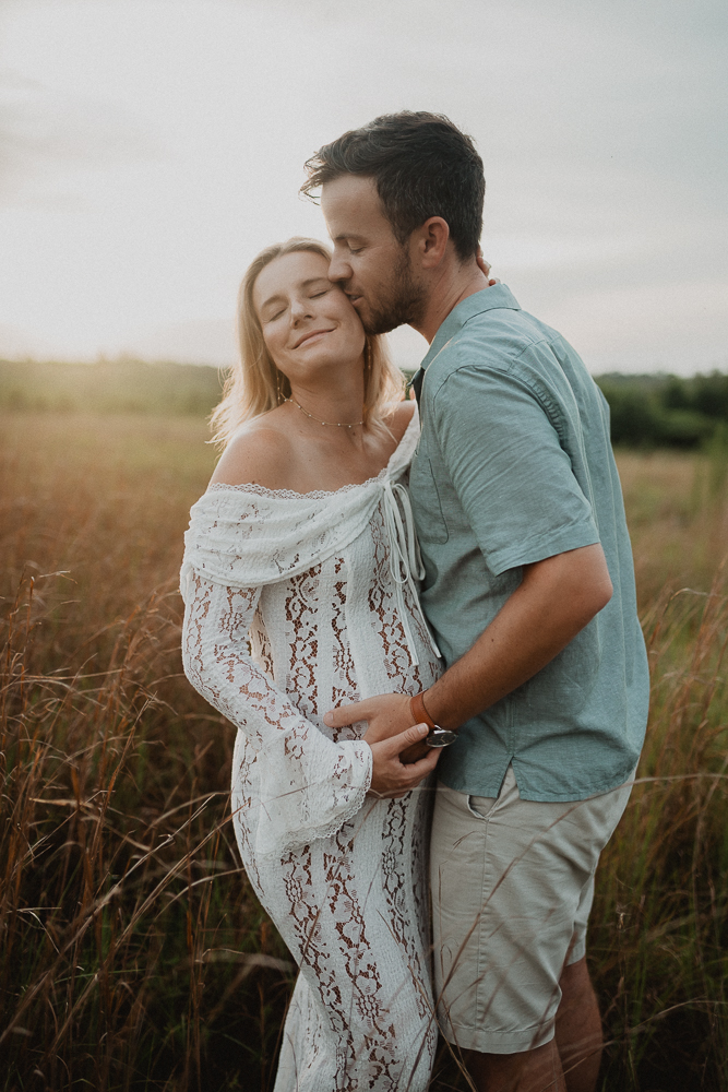 Tender maternity moment of an expecting couple sharing a kiss in a field at sunset, with warm, glowing light outlining the mother's lace gown. The intimate, peaceful vibe reflects the calming prenatal sessions offered by Lucky Elephant Yoga and Wellness. Photo by Victoria Vasilyeva Photography.