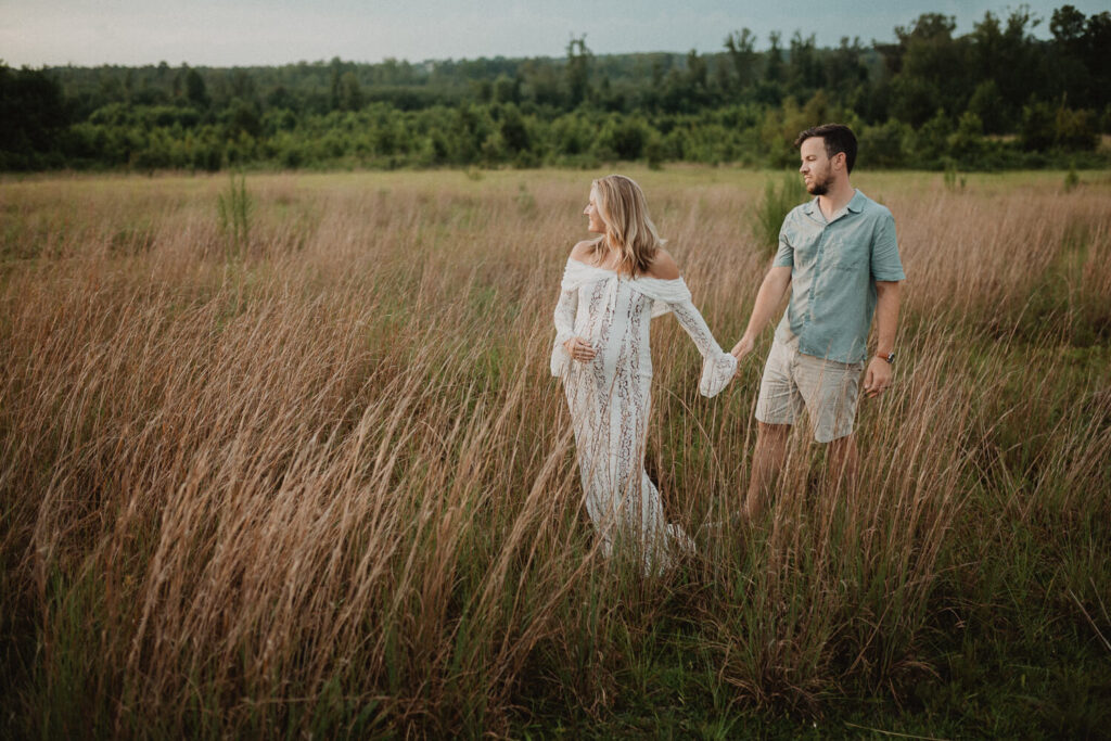 Wide-angle maternity photo of a pregnant woman and her partner walking hand-in-hand through a vast open field with tall grass and soft evening light. Captured by Raleigh maternity photographer Victoria Vasilyeva Photography.