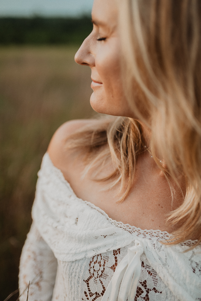 Close-up maternity portrait of an expecting mother in a white lace off-the-shoulder gown, standing in a grassy field at sunset with her eyes closed and soft golden light on her face. Photo by Raleigh maternity photographer Victoria Vasilyeva Photography.