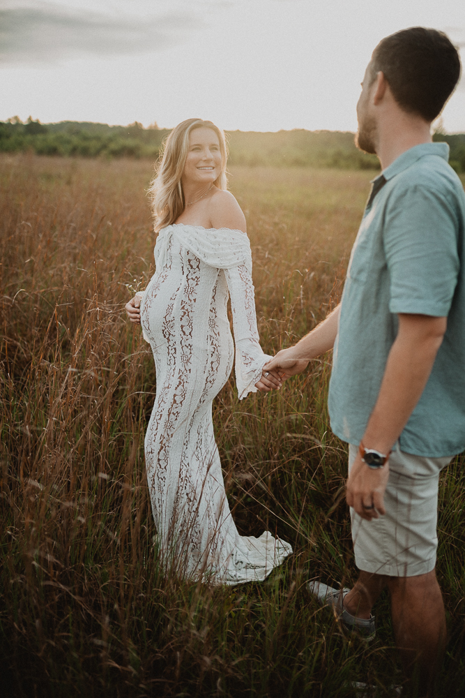 Pregnant woman in a fitted white lace gown smiling at her partner while holding his hand in a tall grass field during golden hour. The warm sunset glow creates a peaceful, connected atmosphere similar to mindful prenatal practices at Lucky Elephant Yoga and Wellness. Captured by Victoria Vasilyeva Photography.