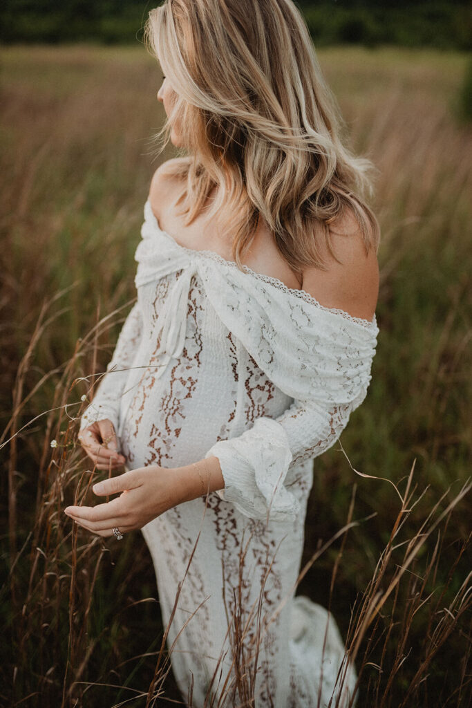 Expecting mother standing in tall grass, gently touching the wildflowers around her while wearing a delicate white lace maternity dress. Photo by Victoria Vasilyeva Photography.