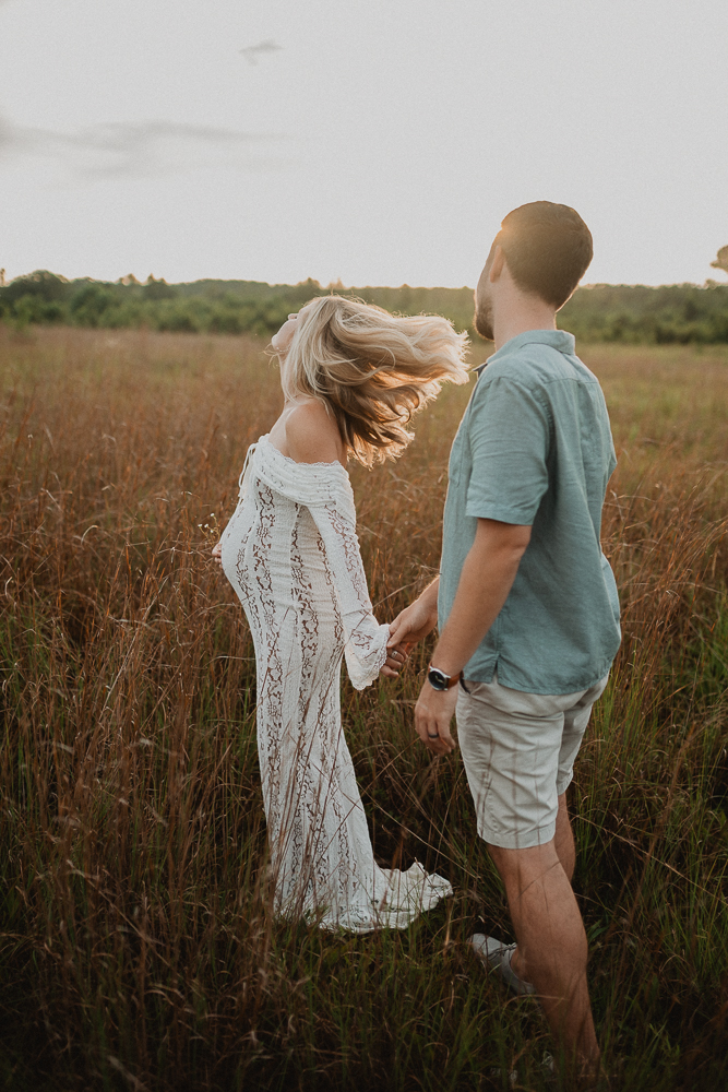 Pregnant woman walking through a field with her partner, her hair blowing in the wind as she wears a long white lace gown after the yoga class at Lucky Elephant Yoga and Wellness. Captured by Raleigh maternity photographer Victoria Vasilyeva Photography.