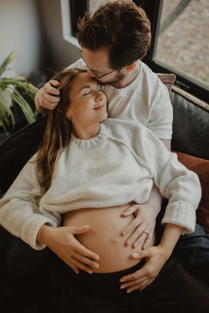 Pregnant woman relaxing at home while embraced by her partner, highlighting prenatal wellness and comfort associated with Moonshadow Medical Massage prenatal care.