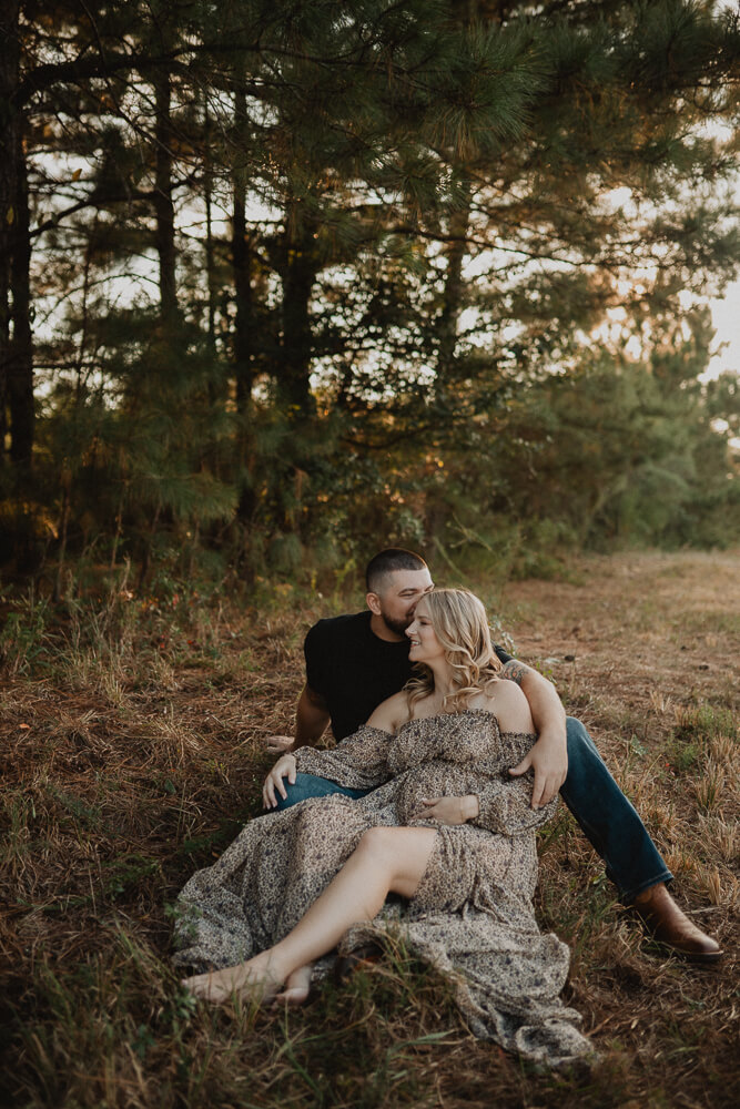 Expecting couple sitting together in a pine forest near Natural Beginnings Birth Center at sunset, sharing a quiet moment during their maternity session. Photographed by Victoria Vasilyeva Photography.