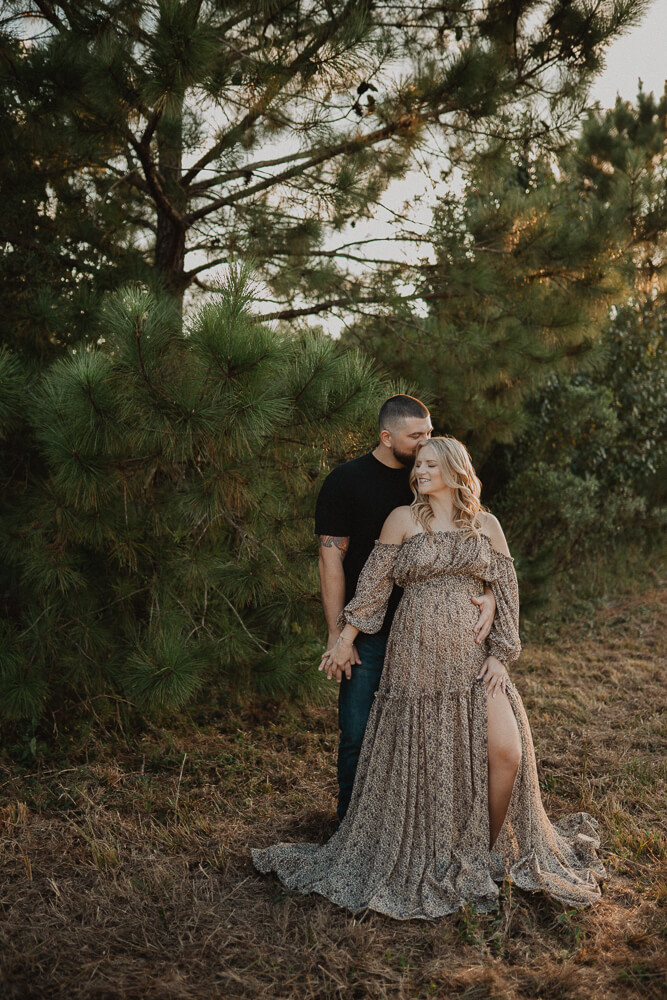 Expecting couple standing together under tall pine trees, holding hands during their maternity session. Maternity photo session with Victoria Vasilyeva Photography.