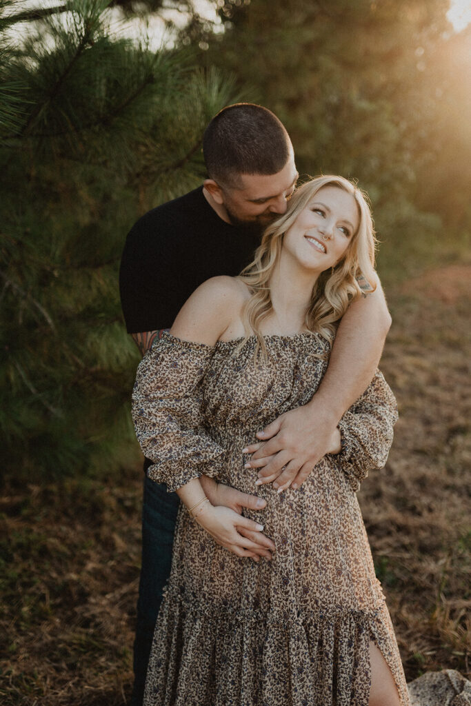 Pregnant woman in maxi floral dress smiling while her partner in black t-shirt hugs her from behind during a golden-hour maternity session. Image by Victoria Vasilyeva Photography.