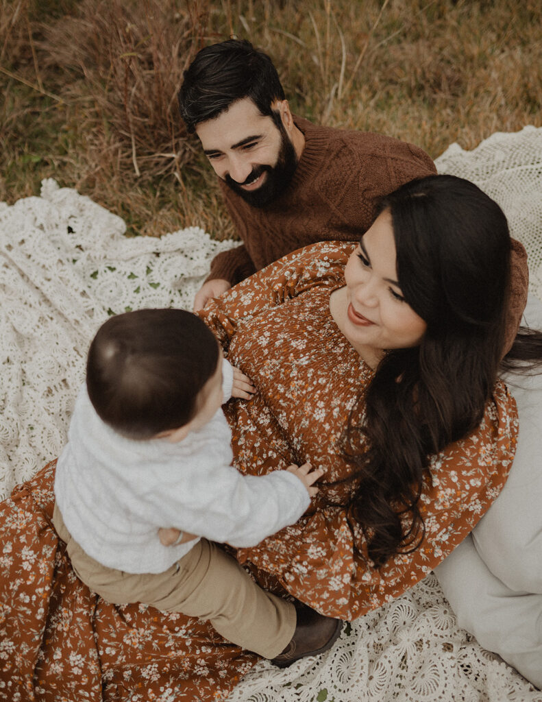 Family sitting together on a blanket in a grassy field, smiling at their son during an outdoor session. Photo used in a blog featuring Over the Moon Play Space.