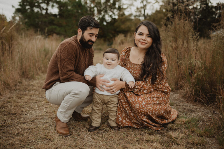 Parents kneeling beside their toddler son in a natural field setting, smiling during a family session after visiting Over the Moon Play Space.
