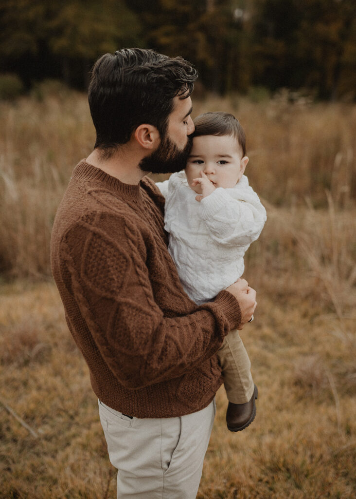 Father holding his baby close and kissing him while standing in a field during a cozy outdoor family session with Victoria Vasilyeva Photography.