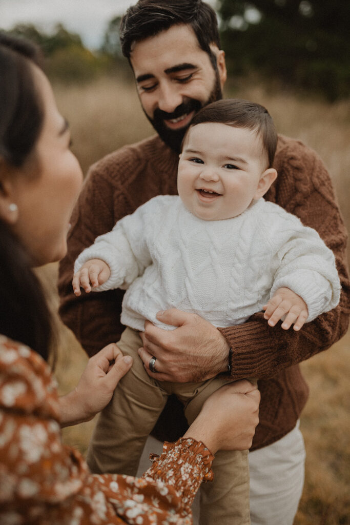 Laughing baby boy in white cozy sweater held by both parents during an outdoor fall session, creating a warm family moment.