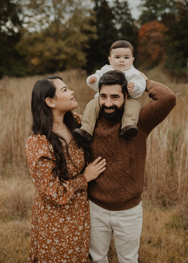Parents smiling as their baby sits on the father’s shoulders during a family photo session in a tall grassy field. Family photo session near Over the Moon Play Space with Raleigh family photographer.