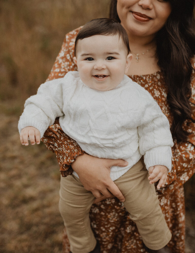 Close-up of a smiling baby wearing a white sweater while being held by a mom during a family session. Image used in Over the Moon Play Space article.