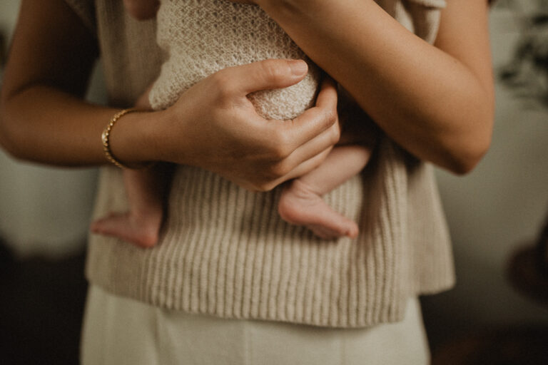 Close-up detail of a newborn’s legs and feet resting in a parent’s arms during a breastfeeding session at home. Photo by Victoria Vasilyeva Photography.