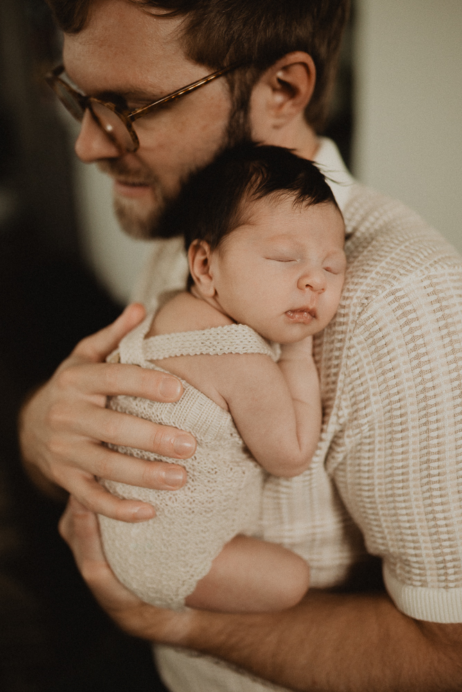 Father gently holding his sleeping newborn girl against his chest in a cozy indoor setting, highlighting family support during early feeding days. Lactation consultant Cary NC, photo by Victoria Vasilyeva Photography.