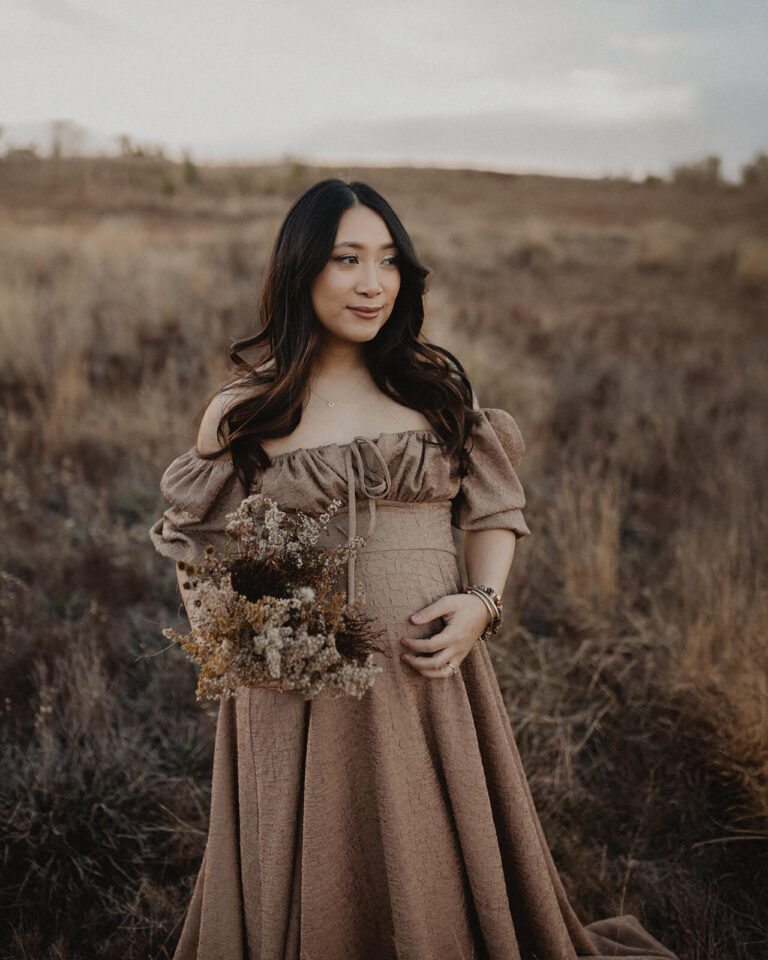 Pregnant woman with black long hair wearing a neutral-toned dress and holding dried florals during an outdoor maternity session in a field near Raleigh, NC. Maternity photography by Victoria Vasilyeva Photography.