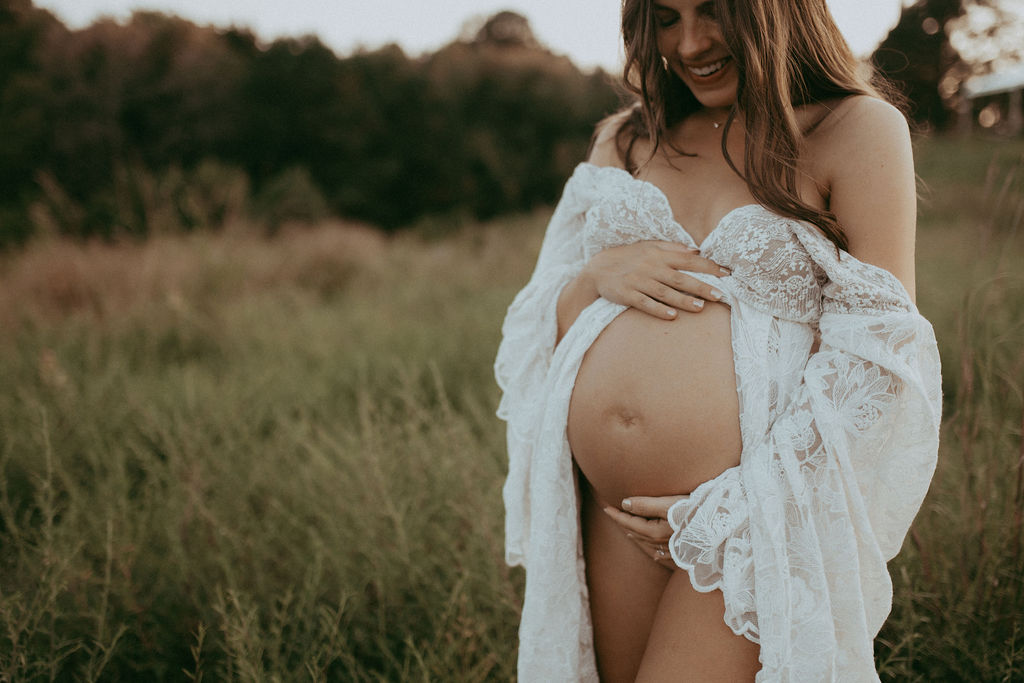 Pregnant woman in a white lace robe holding her bare baby bump while smiling softly during a maternity photo session at NCMA, surrounded by tall grass and soft natural light.