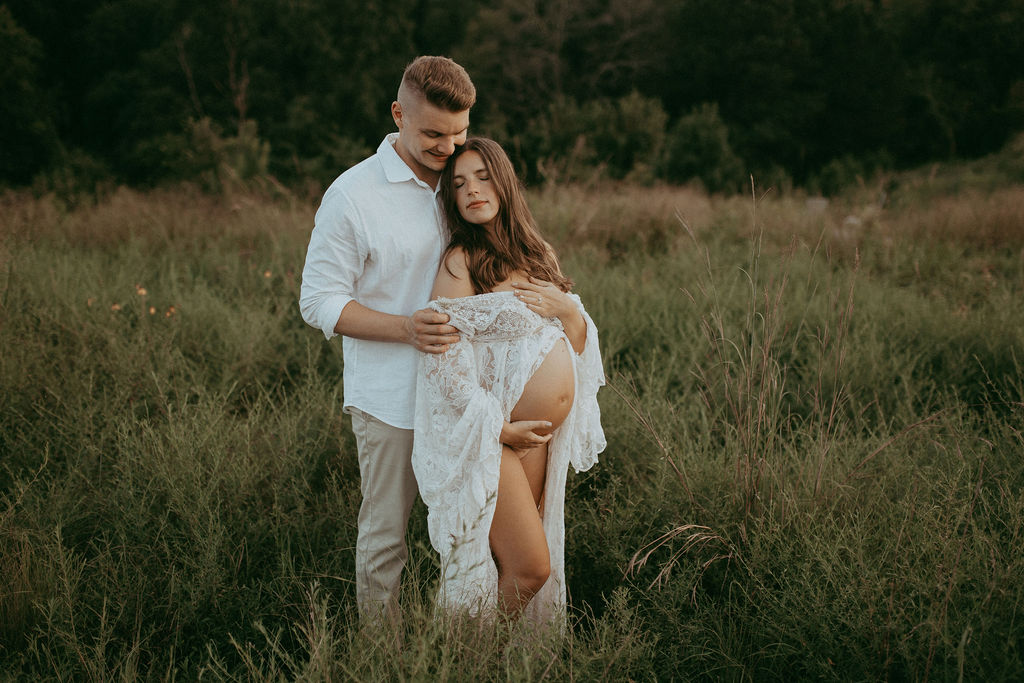 Expecting couple embracing in a grassy field, the woman wearing a flowing white lace gown from Reclamation brand showcasing her baby bump while her partner holds her tenderly during their maternity photoshoot at NCMA in Raleigh.