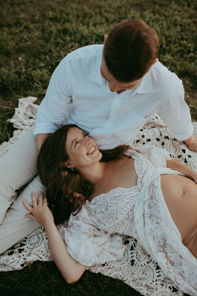 Pregnant woman with long dark hair lying on a crochet blanket in a field, gazing up and smiling at her partner during a relaxed and romantic maternity photo session at NCMA in Raleigh.