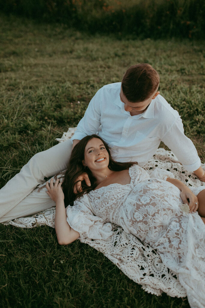 Expecting mother reclining on a blanket with her partner beside her, wearing a detailed white lace gown and smiling warmly during their maternity photo session in Raleigh, NC.