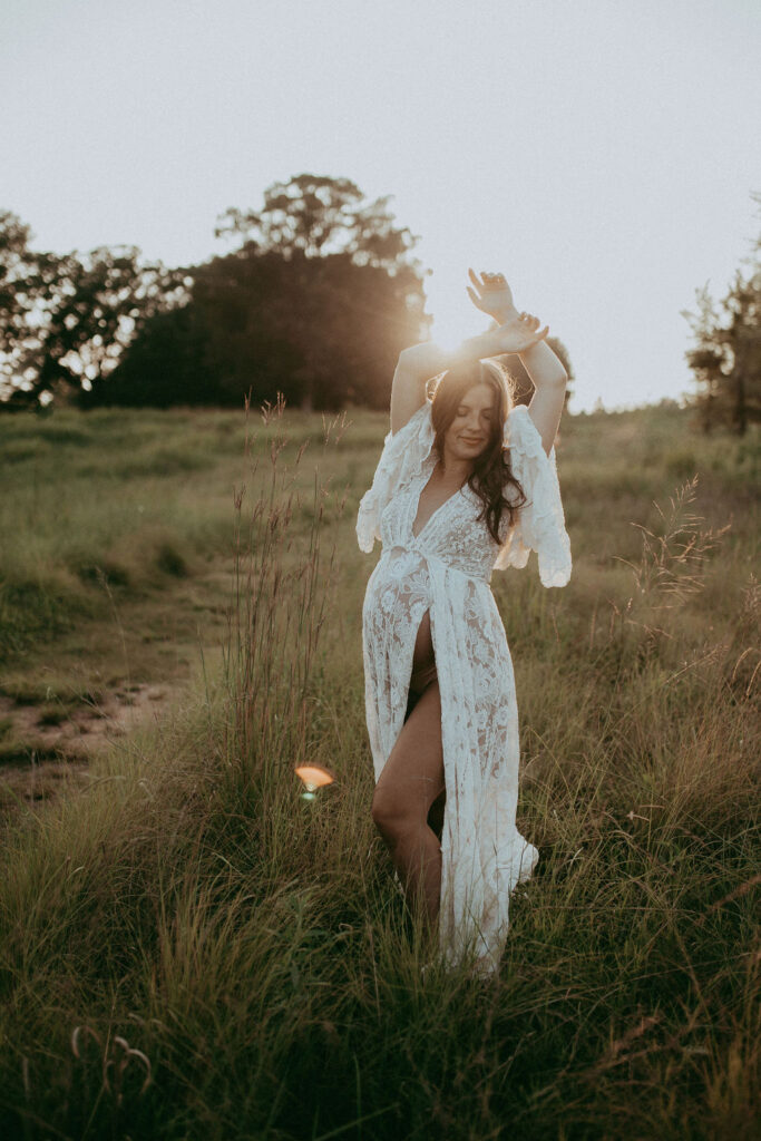 Pregnant woman with dark wavy hair standing in tall grass with arms raised, wearing a white lace gown and glowing in golden-hour sunlight during a dreamy maternity photo session at NCMA.