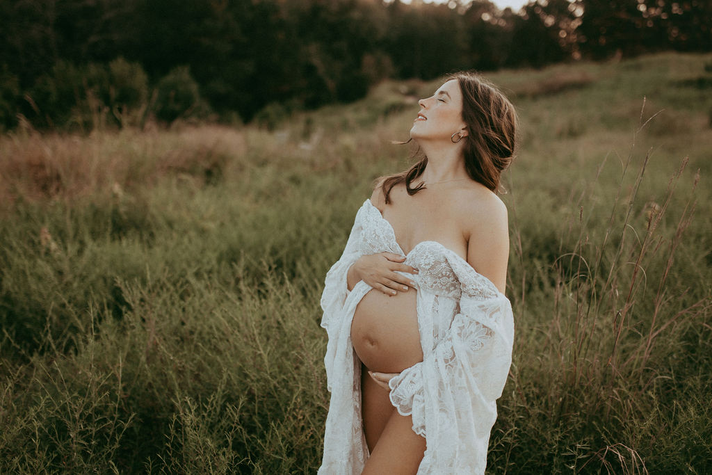 Expecting mother in white lace dress, holding her baby bump and looking up peacefully while standing in a grassy field during a maternity photo session in Raleigh, NC