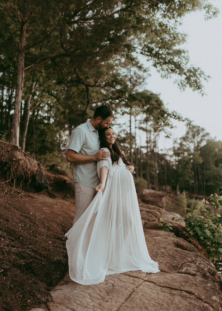 Artistic maternity portrait of an expecting couple posing on a rocky shoreline with forest in the background.