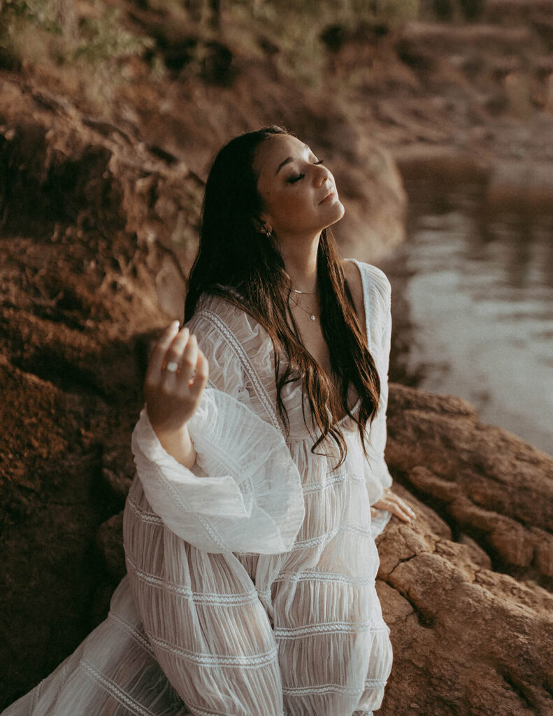 Pregnant woman seated on rocks near the water with eyes closed, enjoying a peaceful moment during an outdoor maternity photography session in Cary NC by Victoria Vasilyeva Photography.