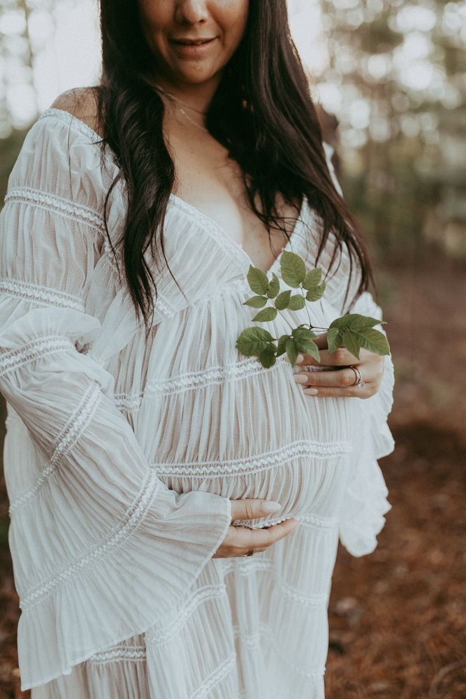 Expecting mother in white maxi dress holding a small green branch while cradling her belly, natural maternity photography Cary NC by Victoria Vasilyeva Photography.