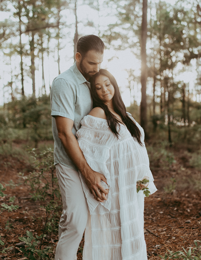 Romantic maternity portrait of a couple standing close in a wooded area with soft evening light, maternity photography Cary NC captured by Victoria Vasilyeva Photography.
