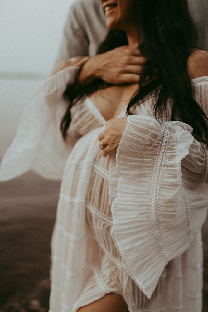 Close-up maternity portrait of an expecting mother smiling while her partner embraces her from behind, wearing a flowing white dress during a lakeside maternity photography session in Cary NC by Victoria Vasilyeva Photography.