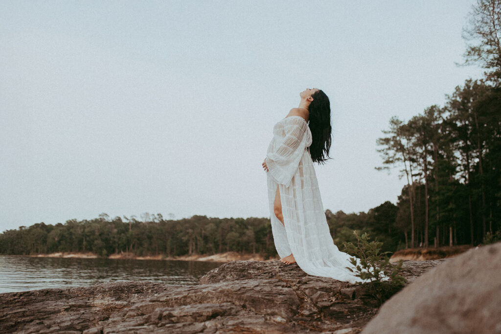 Wide maternity portrait of a pregnant woman standing confidently on a rocky shoreline with water and forest behind her, maternity photography Cary NC by Victoria Vasilyeva Photography.