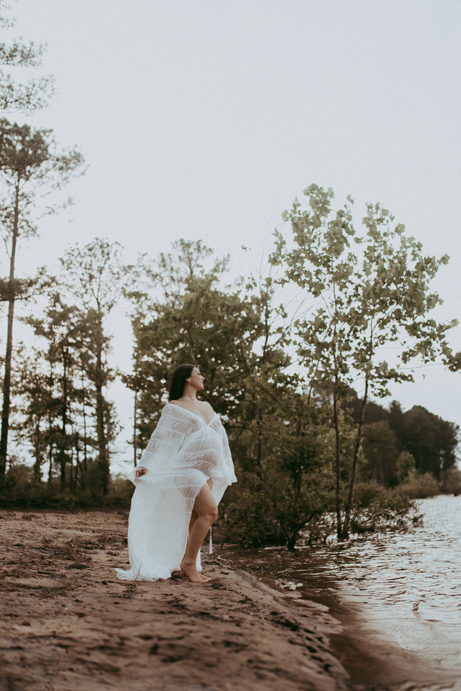 Maternity portrait of an expecting mother walking along a sandy lakeshore with trees in the background, natural maternity photography Cary NC by Victoria Vasilyeva Photography.
