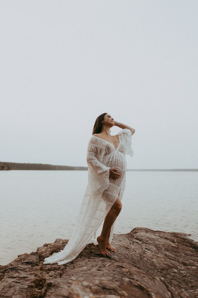 Full-body maternity portrait of an expecting mother in white flowy dress standing barefoot on a rock by the lake in a sheer white dress, maternity photography Cary NC by Victoria Vasilyeva Photography.