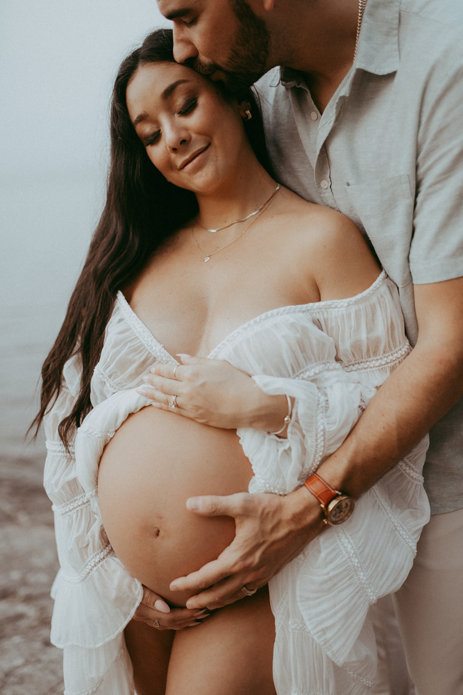 Pregnant woman embraced by her partner during a lakeside maternity session, emotional maternity photography Cary NC photographed by Victoria Vasilyeva Photography.