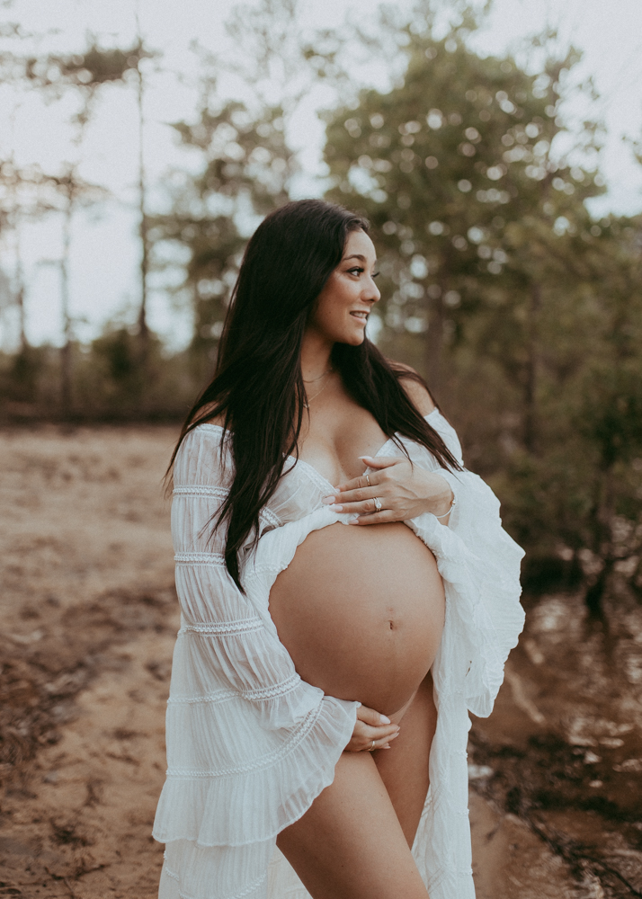 Close-up maternity portrait of a pregnant woman in white dress gently holding her bare baby bump in a natural outdoor setting, maternity photography Cary NC by Victoria Vasilyeva Photography.