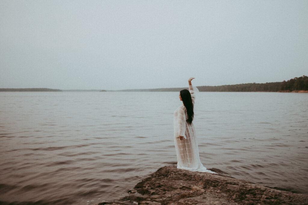 Expecting mother raising her arm toward the sky while standing on a rock by the water, artistic maternity photography in Cary NC captured by Victoria Vasilyeva Photography.
