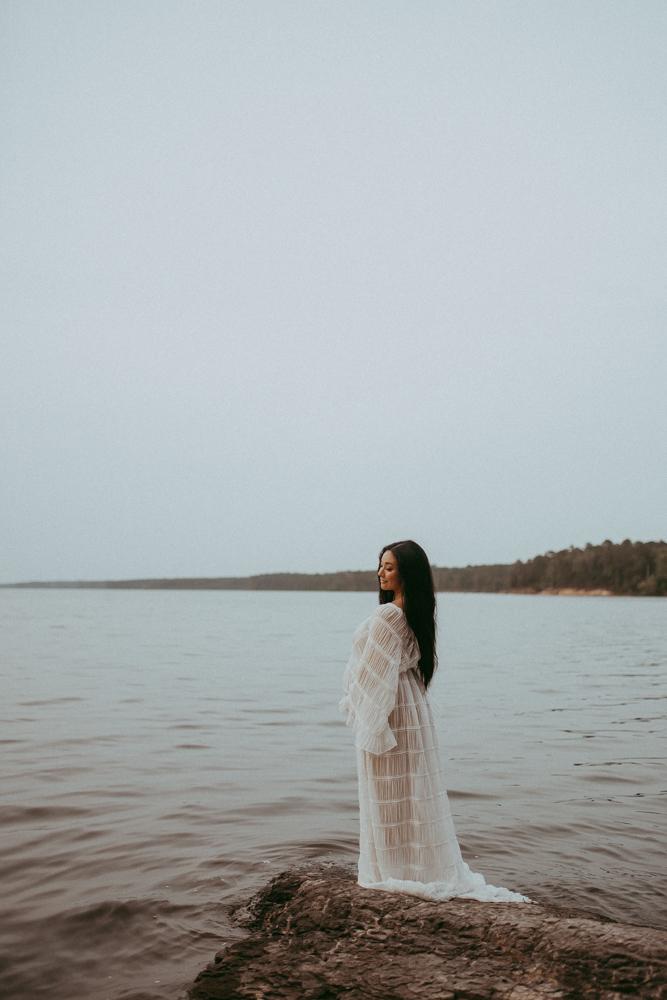 Pregnant woman standing on a rocky lakeshore wearing a flowing white maternity gown, peaceful outdoor maternity photography session in Cary NC by Victoria Vasilyeva Photography.