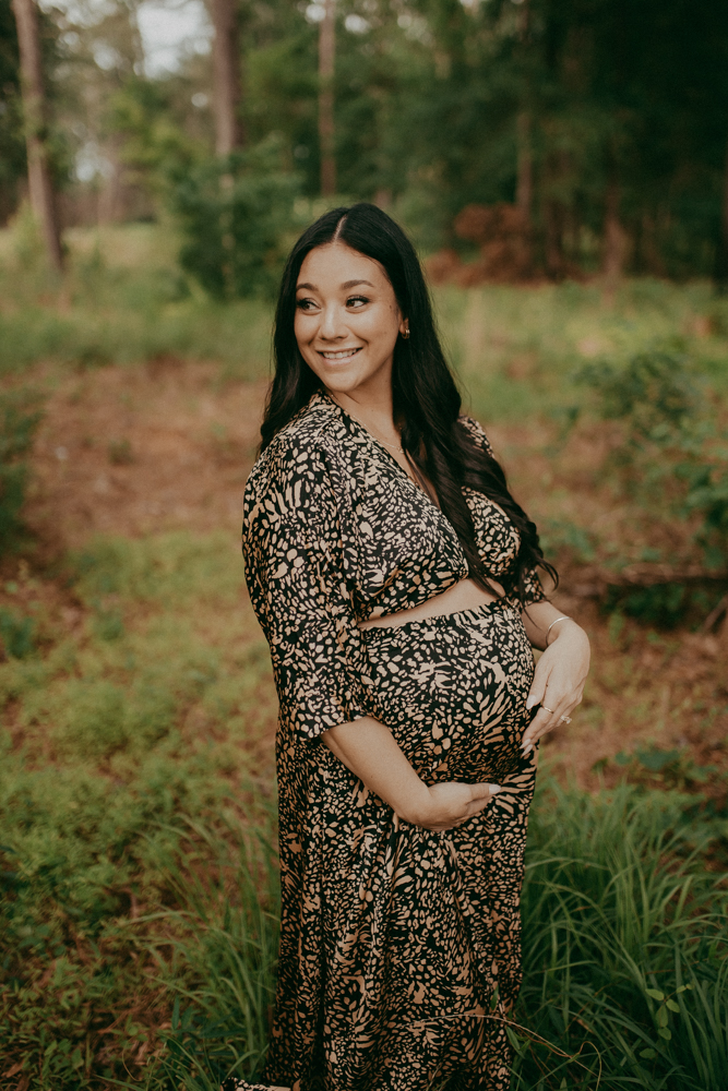 Smiling pregnant woman with long black hair posing in a green forest during golden hour, wearing a patterned maternity outfit and holding her belly. Maternity photography in Cary, NC by Victoria Vasilyeva Photography.